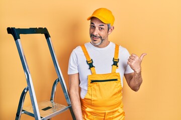 Handsome middle age man with grey hair holding ladder smiling with happy face looking and pointing to the side with thumb up.