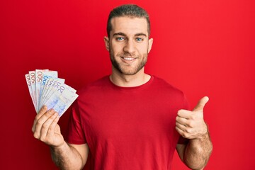 Young caucasian man holding swedish krona banknotes smiling happy and positive, thumb up doing excellent and approval sign