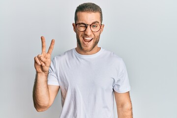 Young caucasian man wearing casual clothes and glasses smiling with happy face winking at the camera doing victory sign. number two.