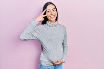 Beautiful woman with blue eyes expecting a baby, touching pregnant belly doing peace symbol with fingers over face, smiling cheerful showing victory