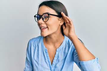 Young hispanic girl wearing casual clothes and glasses smiling with hand over ear listening and hearing to rumor or gossip. deafness concept.