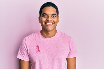 Young handsome hispanic man wearing pink cancer ribbon on shirt looking positive and happy standing...