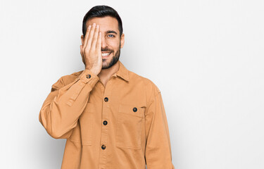 Young hispanic man wearing casual clothes covering one eye with hand, confident smile on face and surprise emotion.