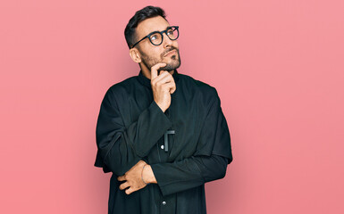 Young hispanic man wearing priest uniform serious face thinking about question with hand on chin, thoughtful about confusing idea