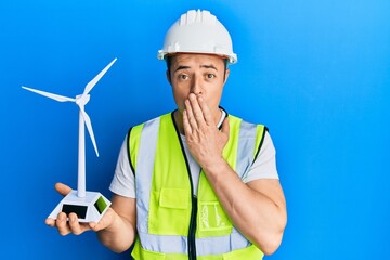 Handsome young man holding solar windmill for renewable electricity covering mouth with hand, shocked and afraid for mistake. surprised expression