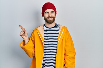 Caucasian man with beard wearing yellow raincoat with a big smile on face, pointing with hand finger to the side looking at the camera.