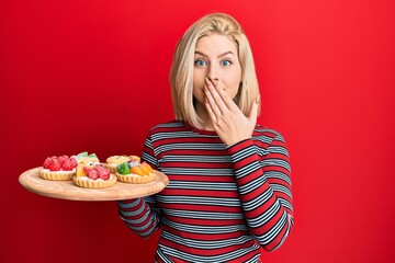 Young blonde woman holding sweet pastries covering mouth with hand, shocked and afraid for mistake. surprised expression