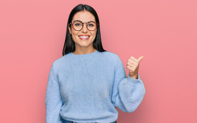 Young hispanic woman wearing casual clothes and glasses smiling with happy face looking and pointing to the side with thumb up.