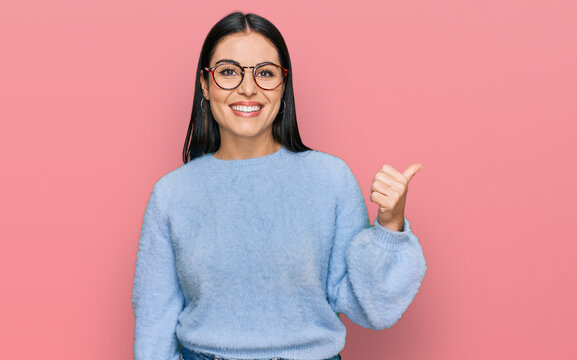 Young hispanic woman wearing casual clothes and glasses smiling with happy face looking and pointing to the side with thumb up.