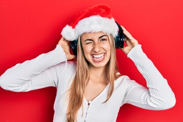 Beautiful hispanic woman wearing christmas hat and headphones winking looking at the camera with sexy expression, cheerful and happy face.