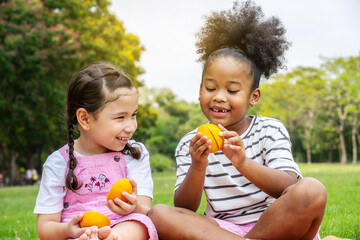 Two African American little girl sitting on the mat and eating oranges fruits in the autumn park ,...