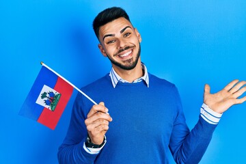 Young hispanic man with beard holding haiti flag celebrating achievement with happy smile and winner expression with raised hand