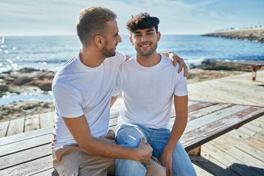 Young gay couple smiling happy sitting on the bench at the beach promenade.