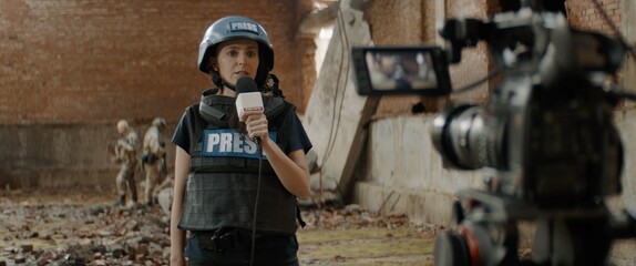 Behind the scenes of female war journalist correspondent wearing bulletproof vest and helmet reporting live near destroyed building, military personnel in the background - obrazy, fototapety, plakaty
