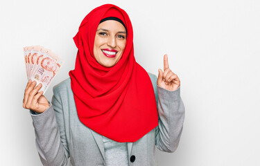 Beautiful hispanic woman wearing islamic hijab holding singapore dollars banknotes surprised with an idea or question pointing finger with happy face, number one