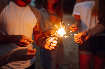 Group of people holding sparklers at party on the beach. Young friends have fun with fire sparkles at picnic at sunset. Summer holidays, vacation, relax and lifestyle concept.