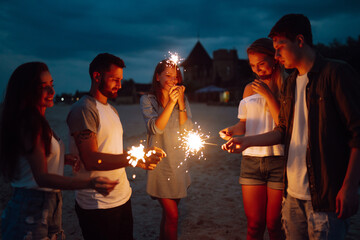Group of people holding sparklers at party on the beach. Young friends have fun with fire sparkles at picnic at sunset. Summer holidays, vacation, relax and lifestyle concept.