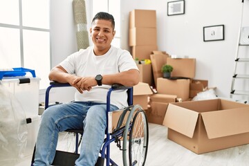 Young hispanic man sitting on wheelchair at new house looking positive and happy standing and...