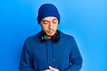 Hispanic young man wearing sweatshirt and headphones with hand on stomach because indigestion,...