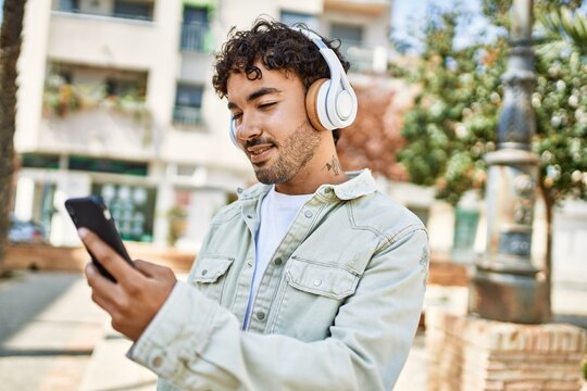 Handsome hispanic man with beard smiling happy outdoors on a sunny day wearing headphones listening to music with smartphone