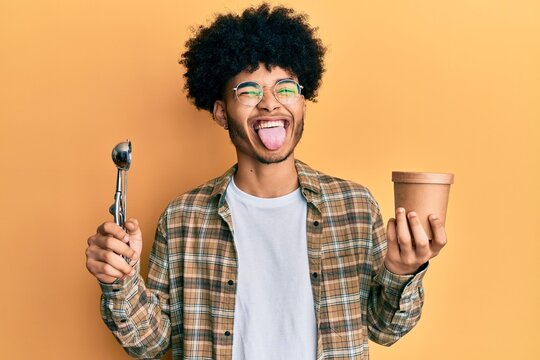 Young african american man with afro hair holding ice cream and ice cream scoop sticking tongue out happy with funny expression.
