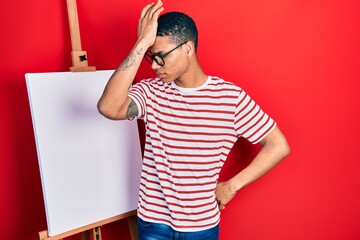 Young african american guy standing by painter easel stand surprised with hand on head for mistake,...