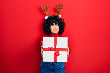 Young middle east woman wearing deer christmas hat holding gift clueless and confused expression. doubt concept.