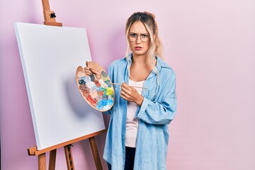 Beautiful young blonde woman standing drawing with palette by painter easel stand skeptic and nervous, frowning upset because of problem. negative person.