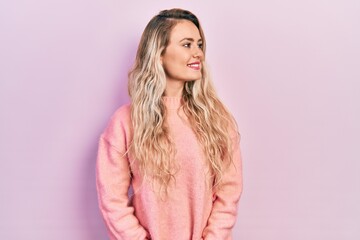 Beautiful young blonde woman wearing pink sweater looking away to side with smile on face, natural expression. laughing confident.