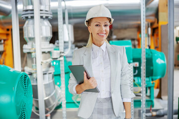 Middle-aged successful blond female supervisor in suit with protective helmet on head standing in power plant with tablet in hands and looking at camera.