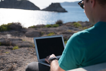 Young man working on a laptop sitting on a bench in a viewpoint in front of the sea. Remote working concept.