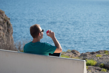 Young man working on a laptop and drinking coffee sitting on a bench in a viewpoint in front of the sea. Remote working concept.