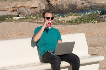 Young man working on a laptop and drinking coffee sitting on a bench in a viewpoint in front of the sea. Remote working concept.