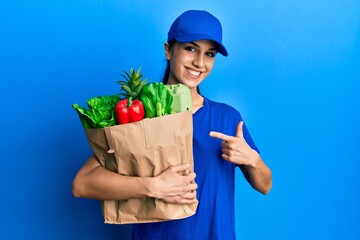Young hispanic woman wearing courier uniform with groceries from supermarket smiling happy pointing...