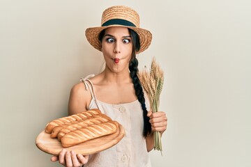 Young hispanic woman holding homemade bread and spike wheat making fish face with mouth and...