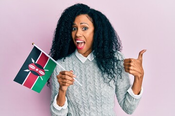 Middle age african american woman holding kenya flag pointing thumb up to the side smiling happy with open mouth