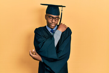 Young african american man wearing graduation cap and ceremony robe hugging oneself happy and...