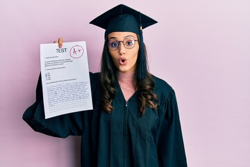 Young hispanic woman wearing graduation uniform holding passed exam scared and amazed with open mouth for surprise, disbelief face
