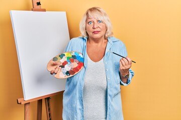 Middle age blonde woman standing drawing with palette by painter easel stand clueless and confused expression. doubt concept.