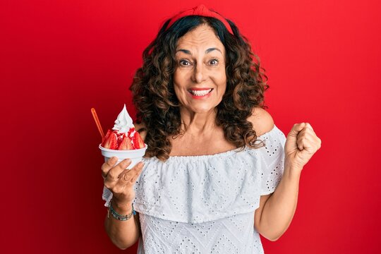 Middle age hispanic woman eating strawberry ice cream screaming proud, celebrating victory and success very excited with raised arm
