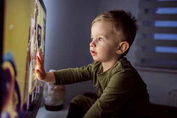 Children's addiction to television and cartoons. A boy touches the TV screen with one hand. Close up of a kid sitting right in front of the TV in his pajamas and staring at a cartoon. Modern parenting - Powered by Adobe