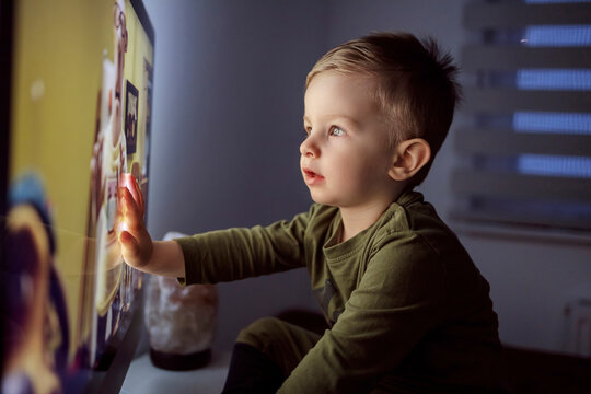 Children's addiction to television and cartoons. A boy touches the TV screen with one hand. Close up of a kid sitting right in front of the TV in his pajamas and staring at a cartoon. Modern parenting