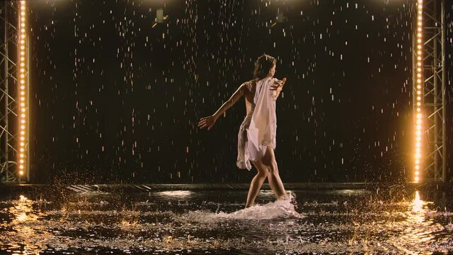 Contemporary sensual ballet choreography performed by slender flexible ballerina amidst the raindrops. Wet woman in beige swimsuit dancing romantically in dark studio. Silhouette. Slow motion.