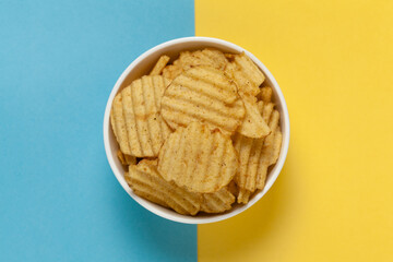 Close up of Wrinkled Wavy Potato Chips in white ceramic bowl, Popular Ready to eat crunchy, salty pale-yellow color over blue yellow background