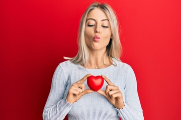 Beautiful blonde woman holding heart making fish face with mouth and squinting eyes, crazy and comical.
