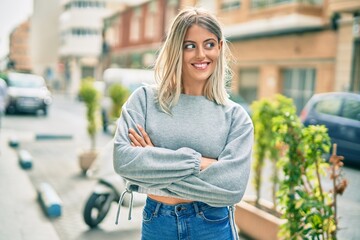 Young blonde girl with arms crossed smiling happy standing at the city.