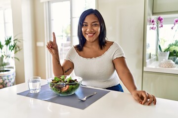 Obraz na płótnie Canvas Young hispanic woman eating healthy salad at home showing and pointing up with finger number one while smiling confident and happy.