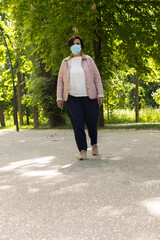 Fototapeta premium Vertical photo with copy space of a middle-aged woman walking with a mask, pink jacket and pink slippers on a sunny day with trees in the background