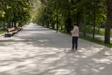 Fototapeta premium Middle-aged woman wearing a coronavirus mask texting on a smart phone while walking through a tree-lined park with a large path in a pink jacket