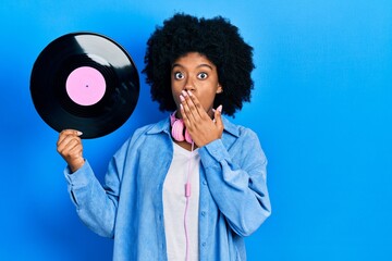 Young african american woman wearing headphones holding vinyl disc covering mouth with hand,...